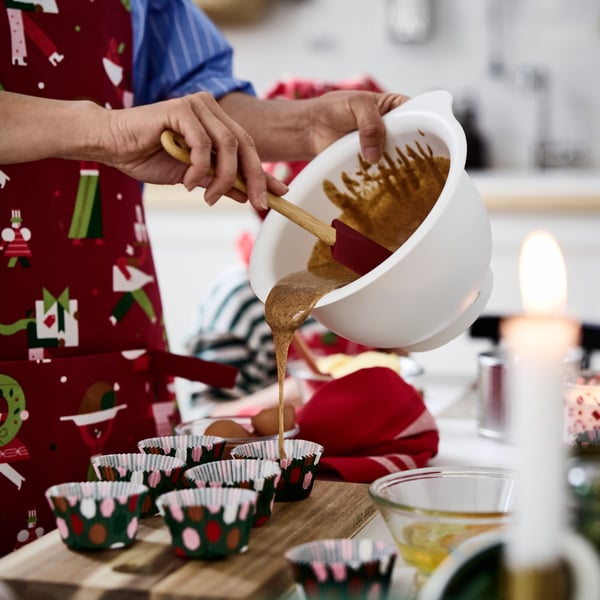 A person wearing an apron pours cake mixture onto festive VINTERFINT baking cups, on a cutting board, on a table.